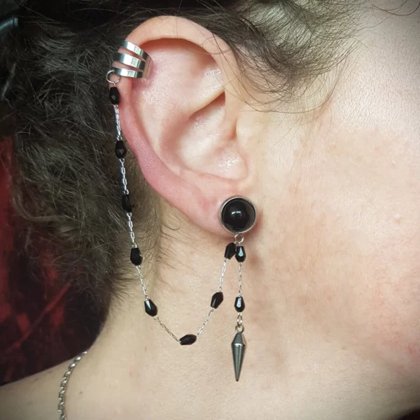 Cette boucle d'oreille chaine et anneau ouvert, pour helix non piercé, se compose d'un clou d'oreille avec onyx accompagné de fines chaines pendantes dont l'une avec pique. Discrète, originale et légère, elle sera parfaite pour les fans du style gothique!